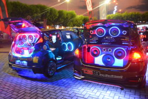 Two custom hatchback cars at a car show with speakers that have RGB lighting around them in the trunk of the cars.