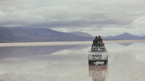 People sitting on top of a vehicle parked in the middle of an open valley with mountains pictured in the distance.