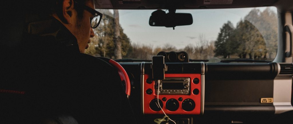 A man wearing glasses glances at his docked phone mounted on his car stereo as he drives through a city.