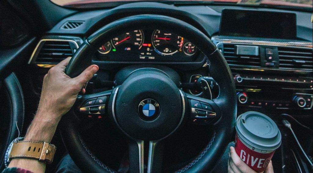 With one hand on the wheel, a driver steers a BMW through a forest road as their passenger leans in with a coffee cup.