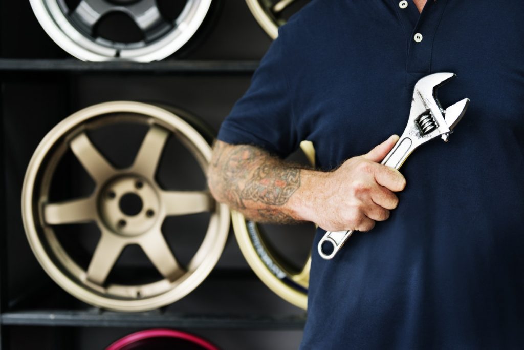 A man with tattoos on his arm holds a crescent wrench as he stands in front of a display of car wheel rims.