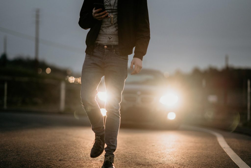 A man in jeans and a leather jacket walks in front of a car, back lit by bright HID headlights.