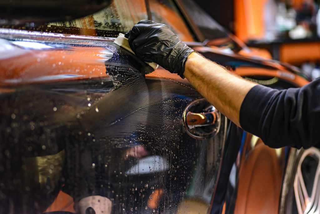 A gloved hand wipes cleaner along a car's body.